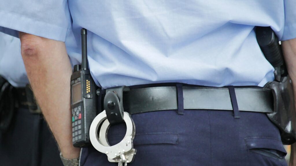 Close-up of a police-officer's belt from behind. We see a walkie-talkie, handcuffs, and a pistol in its holster.
